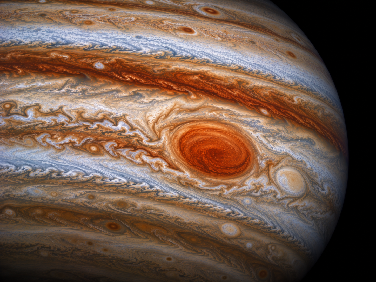 Jupiter’s Great Red Spot appears as a huge rounded storm surrounded by powerful bands of moving atmospheric clouds.